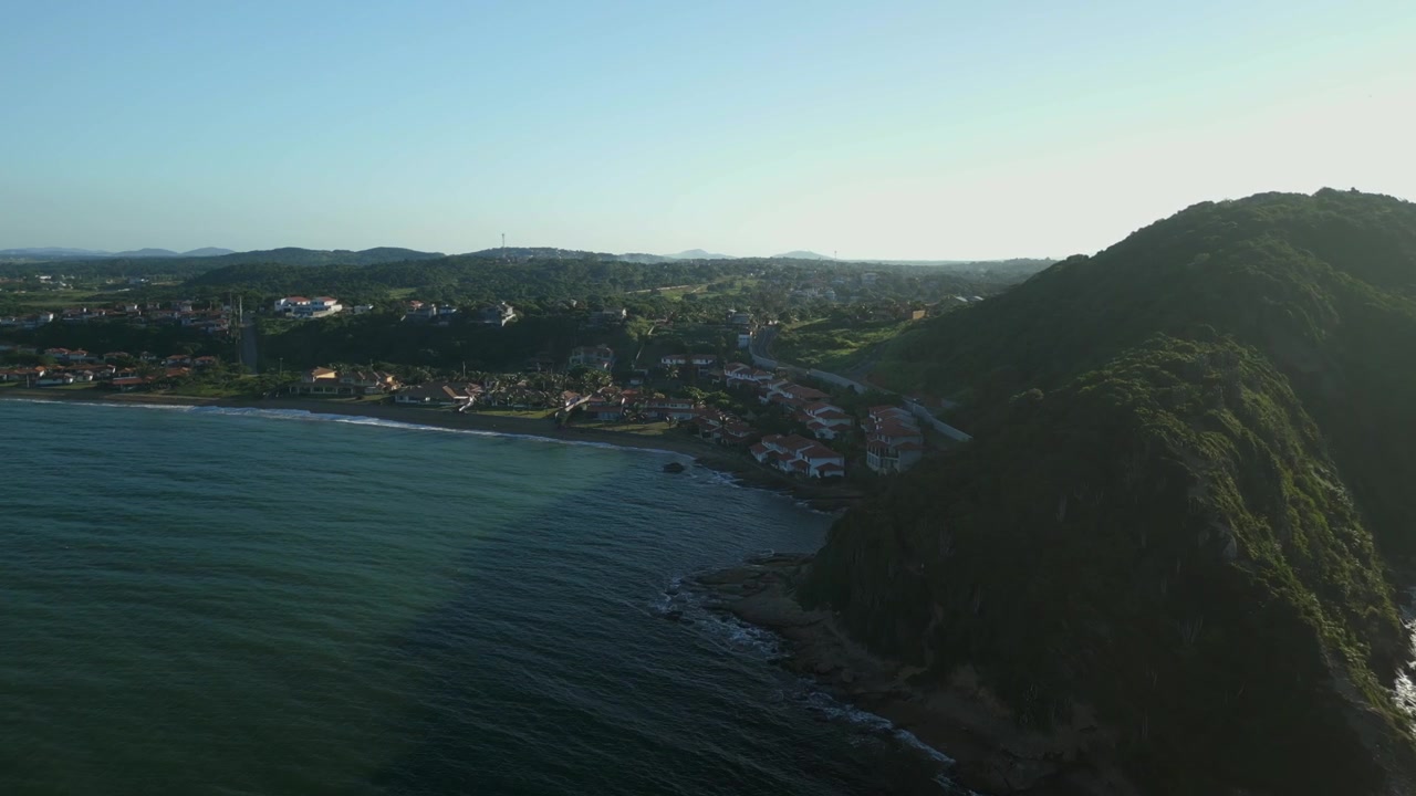 Acque calme del mare a Praia da Marina, Búzios. Oceano e case sulla spiaggia illuminate dai raggi dorati del sole mattutino. Ampia ripresa aerea in planata in 4K a 25 fps.