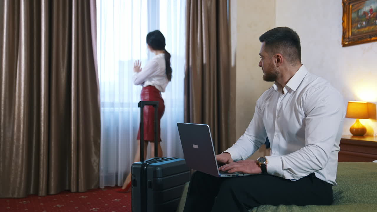 Business colleagues in a hotel room. Handsome man sitting on a bed and working on his laptop. Pretty woman in elegant clothes standing near the window in hotel.
