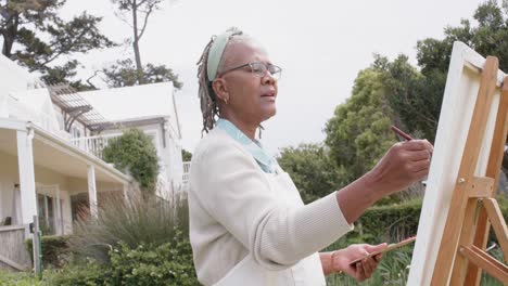 Mujer-Afroamericana-Mayor-Pintando-Sobre-Caballete-De-Madera-En-El-Jardín,-Cámara-Lenta