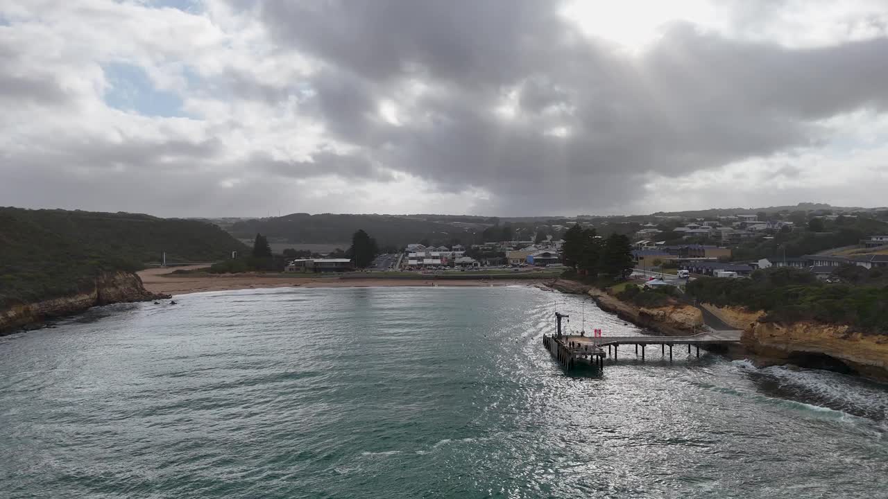 Drone footage captures Port Campbell's coastline with a pier, beach, and town under dramatic skies