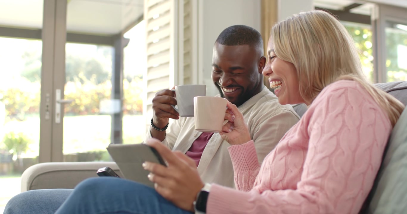 Diverse couple sitting on sofa at home, man raising mug causing woman laughing and scrolling tablet