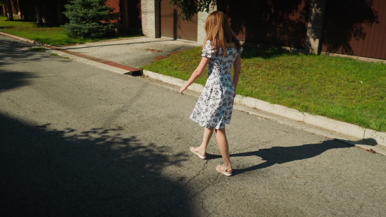 Back view of young woman in floral dress walking thoughtfully down sunny suburban street while butterfly flutters past, casting soft shadow on pavement bordered by green grass and residential fencing