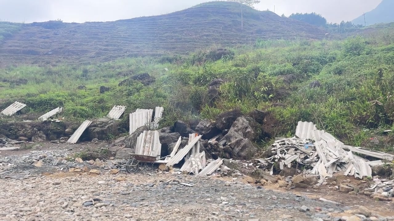 Primo piano roccioso con materiali da costruzione sparsi che conduce verso una collina terrazzata verde sotto un cielo nuvoloso nell'Altopiano Carsico di Dong Van, Vietnam