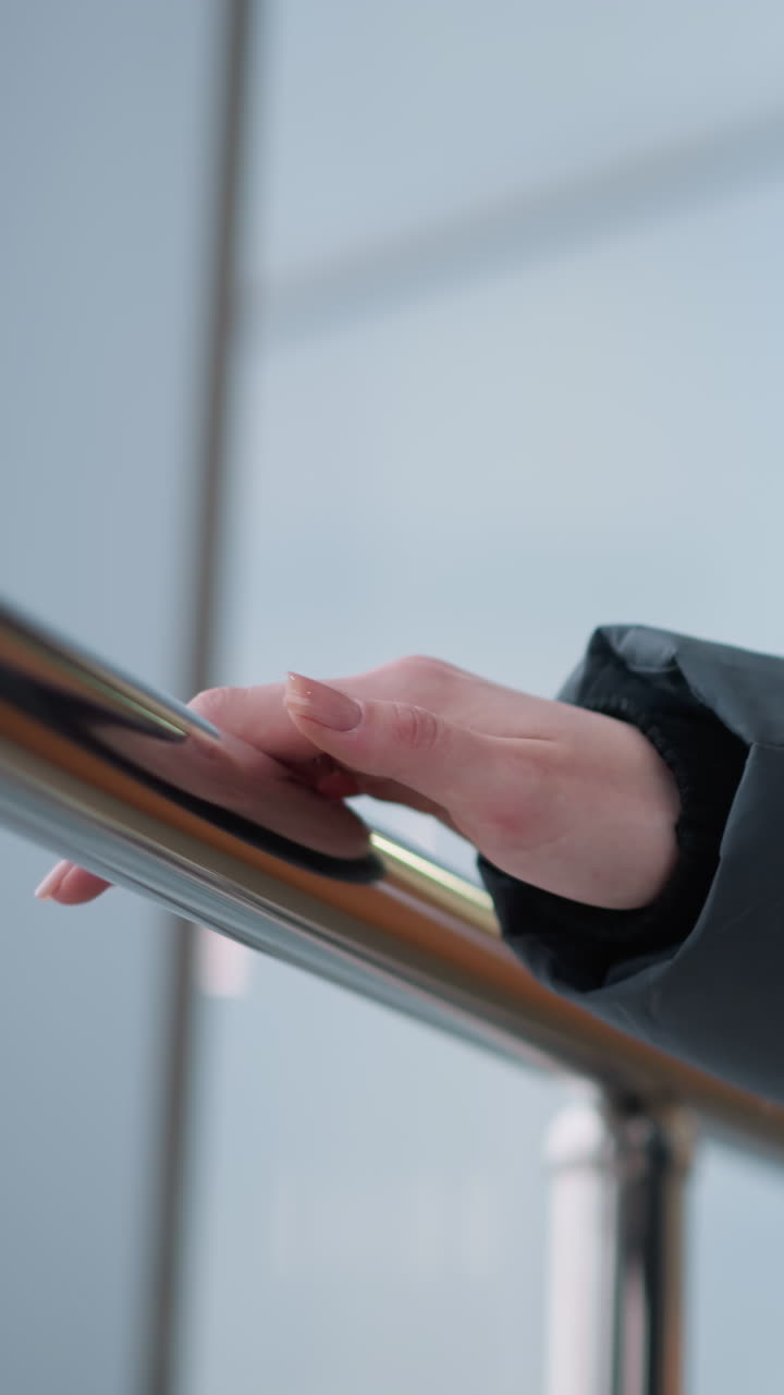 primer plano de la mano de la dama en sudadera negra con dedos adornados con anillos deslizándose en un elegante riel metálico mientras camina, con vidrio de construcción que refleja al aire libre y luz suave que acentúa la superficie lisa