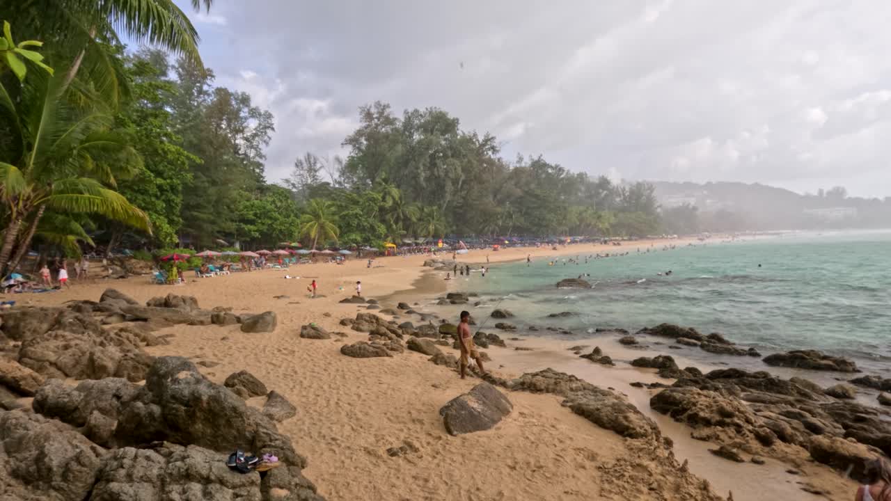 People enjoy a tropical Phuket beach with palm trees, rocky shore, turquoise water, and daylight