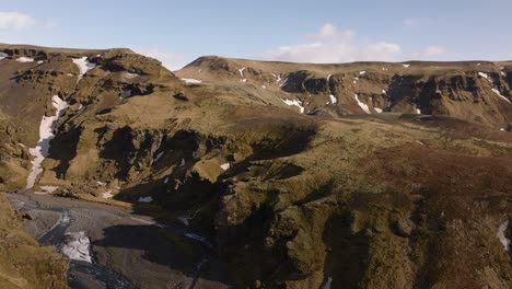 Luftpanoramablick-über-Ein-Gebirgstal-Mit-Einem-Fluss,-Der-Aus-Schmelzendem-Schnee-In-Island-An-Einem-Sonnigen-Morgen-Fließt