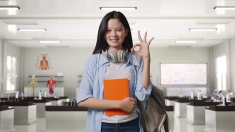 estudiante adolescente asiática con una mochila y algunos libros sonriendo y mostrando un gesto de acuerdo mientras está de pie en el laboratorio de ciencias