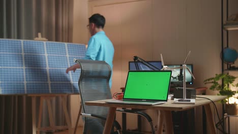 un hombre asiático entra en la oficina mirando la célula solar que está al lado de la pantalla verde de la computadora portátil y la energía eólica en la mesa