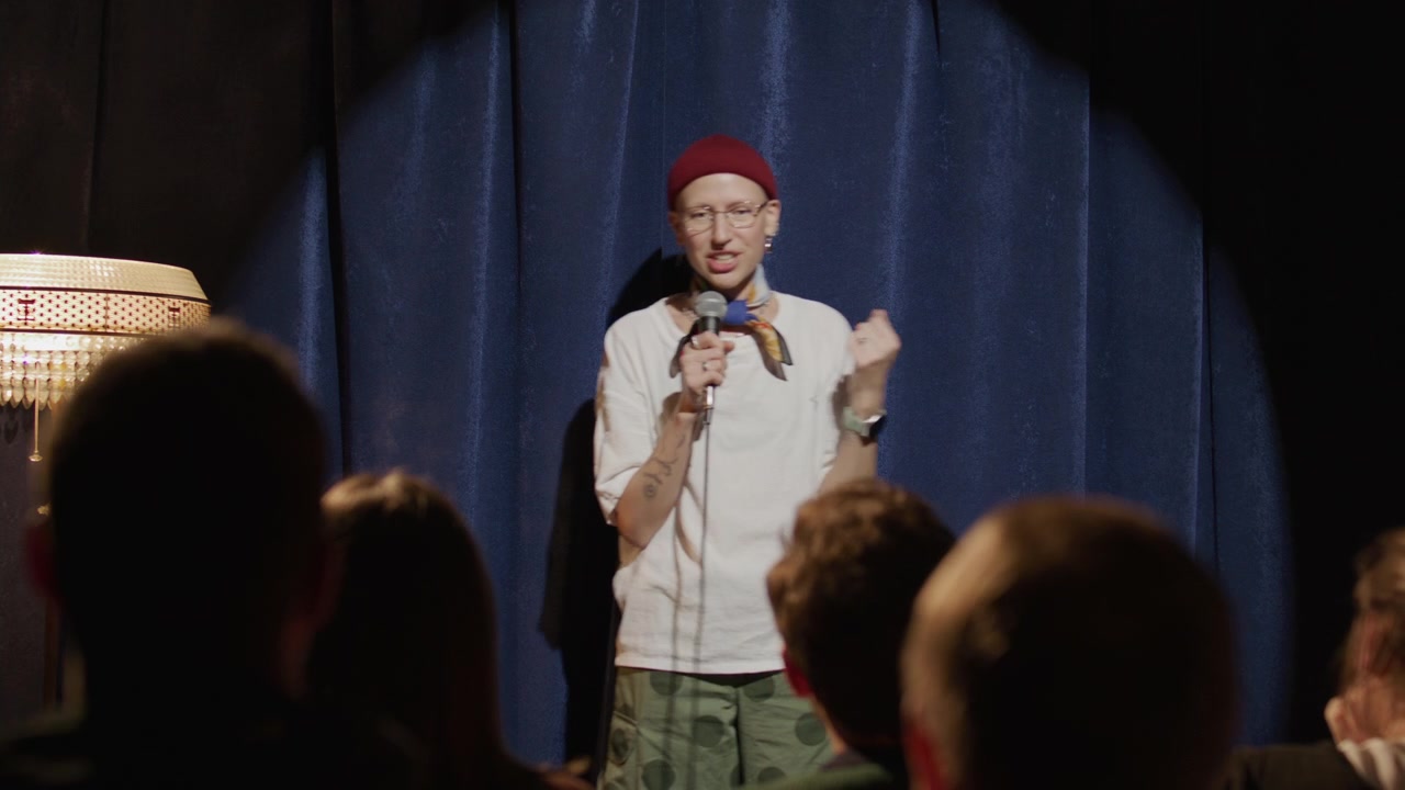 Ung, blid kvinne vinker til publikum og fremfører standup