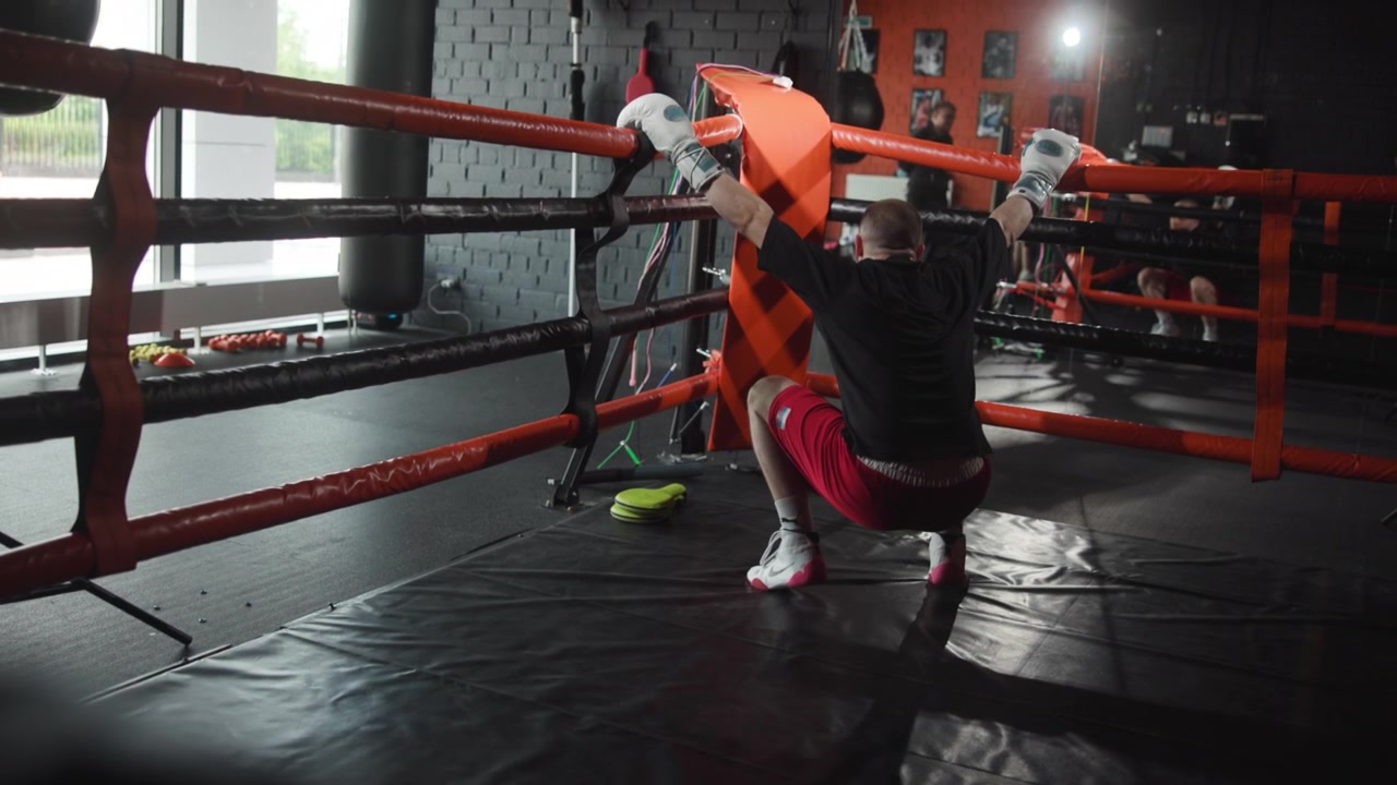 Vista trasera de un boxeador masculino en un ring interior, con las manos enguantadas apoyadas en la cuerda superior y la almohadilla de la esquina mientras se levanta de una posición agachada, con las rodillas dobladas y el peso desplazándose, mostrando recuperación y concentración durante el entrenamiento