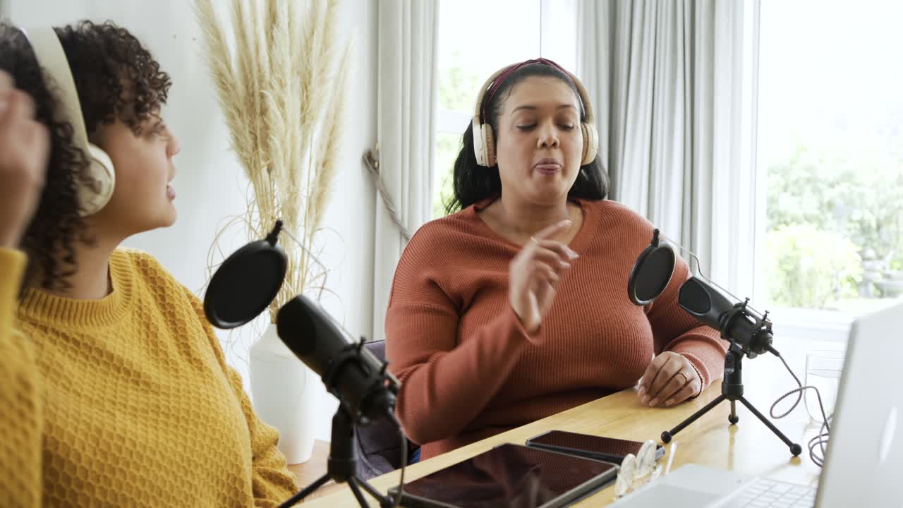 Due co-conduttrici registrano un podcast al tavolo, regolando le cuffie e gesticolando con i microfoni.