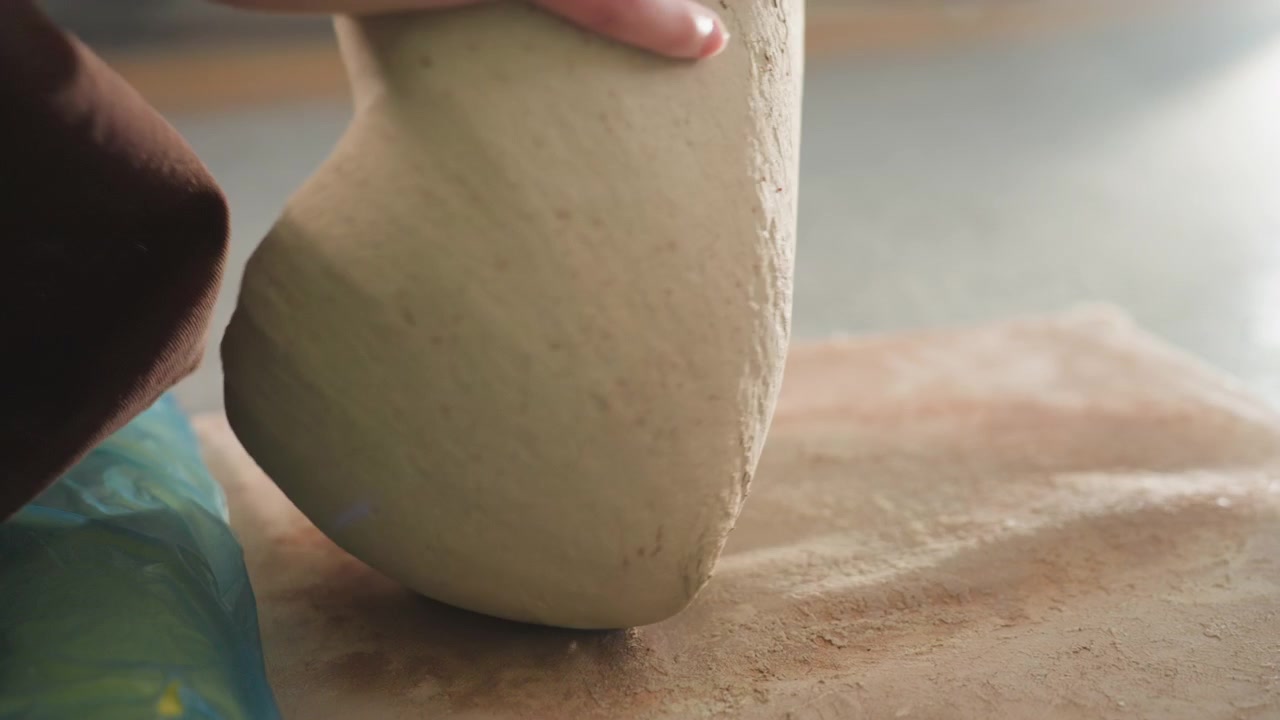 Closeup clay ball on board, pottery workshop prep with quiet focus, artist inspecting surface texture and density, dusted board and mat, earthy tones and soft window light, deliberate stillness