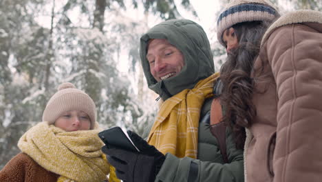 bottom view of a father, mother and daughter dressed in winter clothes lookingt at smartphone in a snowy forest