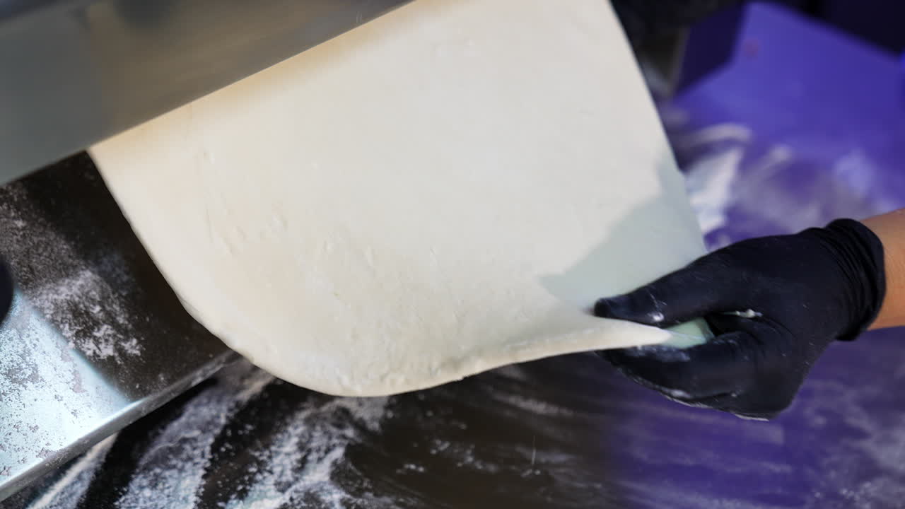Metal roller machine for flattening dough. Gloved hands of a chef pull a flat piece of pastry from a culinary device. Close up.