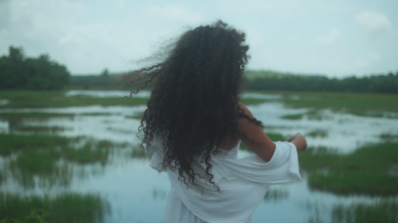 girl with curly hair with her back to the camera looks at the green field meadow on a beautiful day with clouds