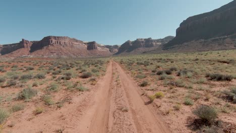 Fpv-Antena-Siguiendo-El-Camino-Sobre-El-Desierto-De-Indian-Creek-Hacia-Enormes-Acantilados-En-Segundo-Plano-En-Un-Día-Soleado