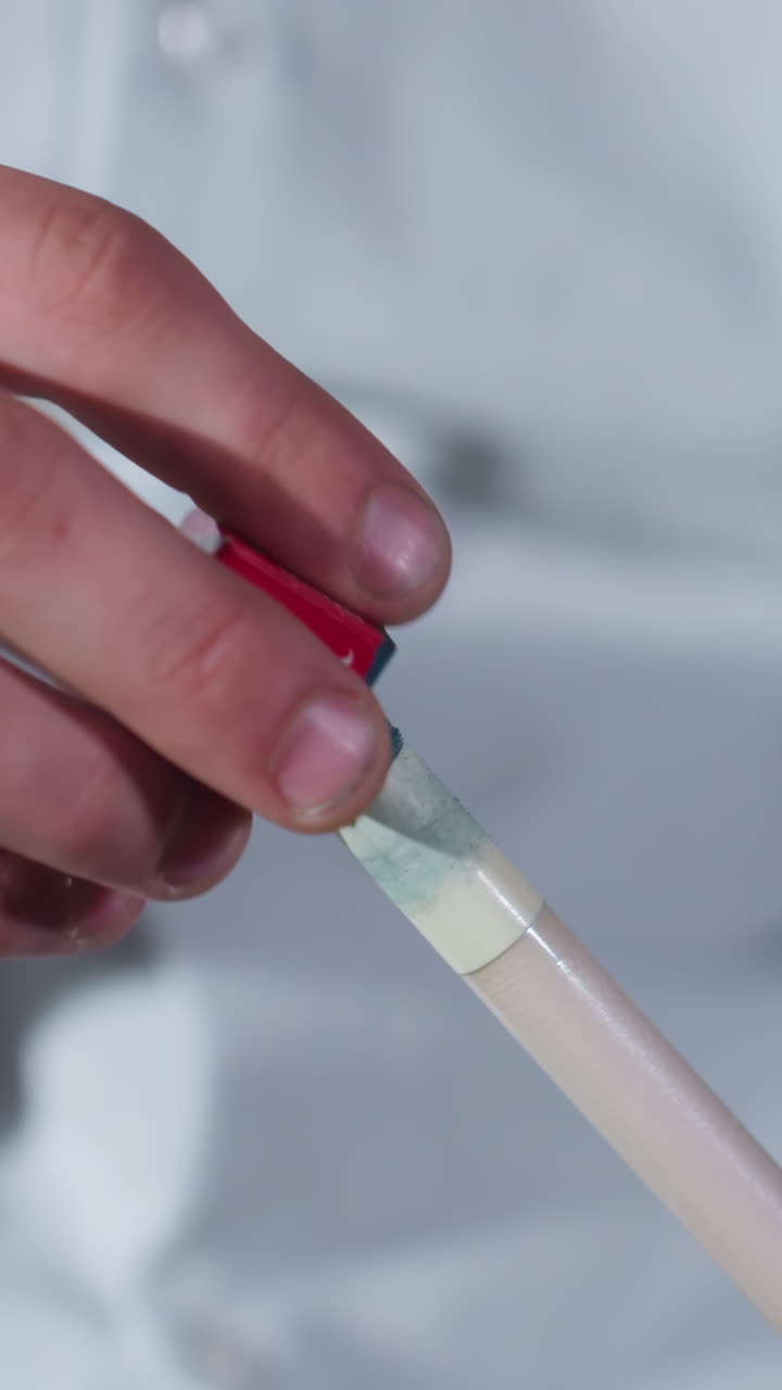 toma de primer plano de la mano del jugador en camisa blanca aplicando cuidadosamente tiza roja y azul al palo de señal antes del juego de billar. el agarre enfocado destaca la precisión, la preparación y la habilidad en el billar