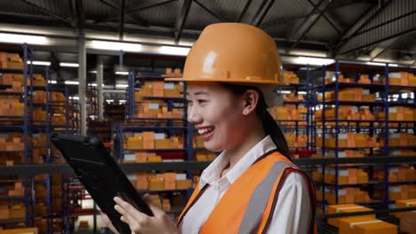 close up side view of asian female engineer with safety helmet working on a tablet while standing in the warehouse with shelves full of delivery goods