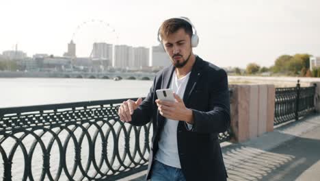 un tipo de raza mixta alegre que usa auriculares caminando al aire libre bailando usando un teléfono inteligente