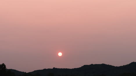 Lapso-De-Tiempo-De-Puesta-De-Sol-Brumoso-Sin-Nubes-Mientras-El-Sol-Va-Detrás-De-Colinas-Boscosas-Distantes,-4k