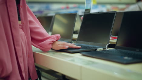 vista del cuerpo de la dama en vestido rosado con bolso negro interactuando con una computadora portátil en exhibición en una tienda de tecnología brillante, la pantalla de la computadora portable está en blanco mientras la opera, con otras computadoras portátiles alineadas