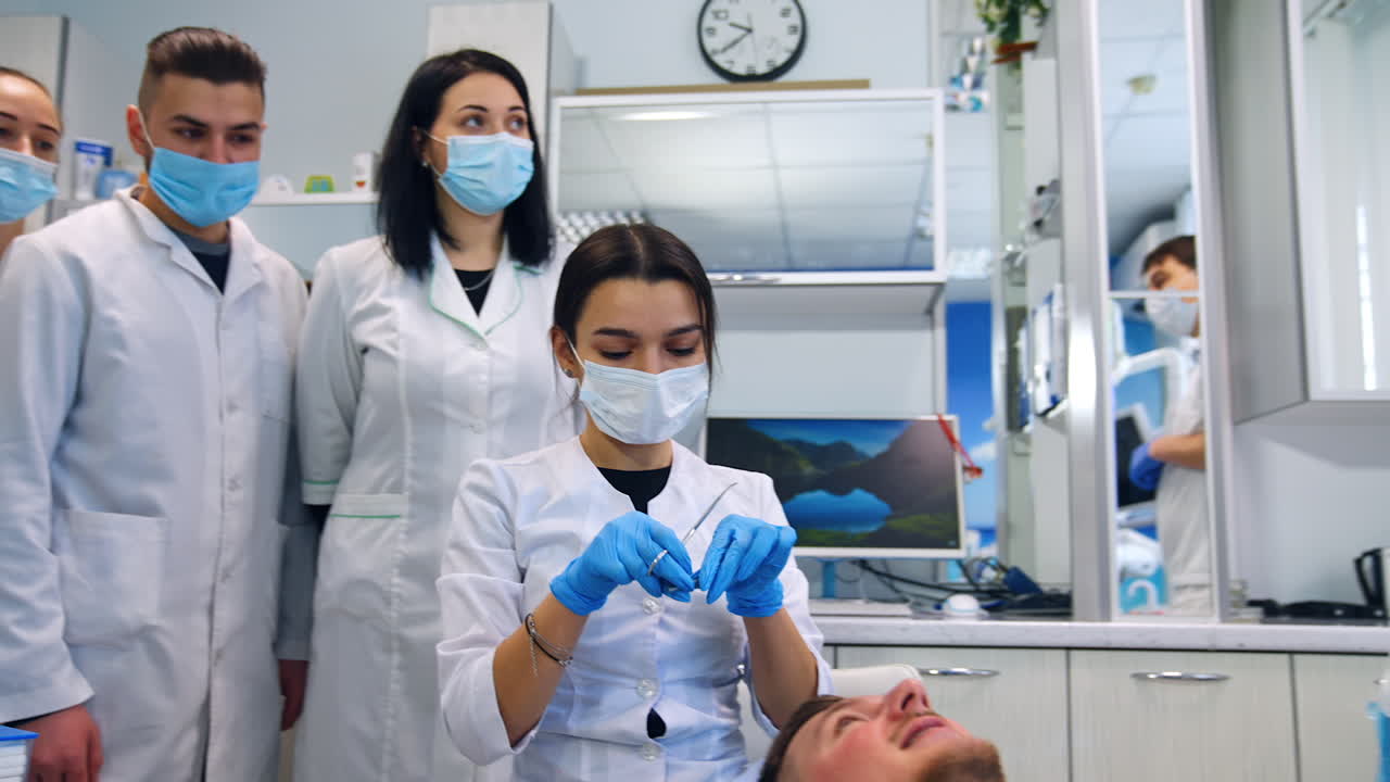 Dentistry department students in masks stand listening to the teacher. Female student sits at the patient.