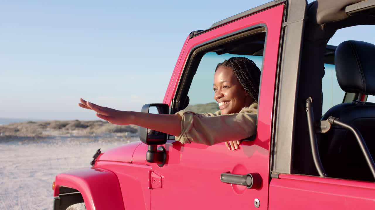 Giovane donna afroamericana si gode una giornata di sole su una jeep rosa vivace durante un viaggio on the road
