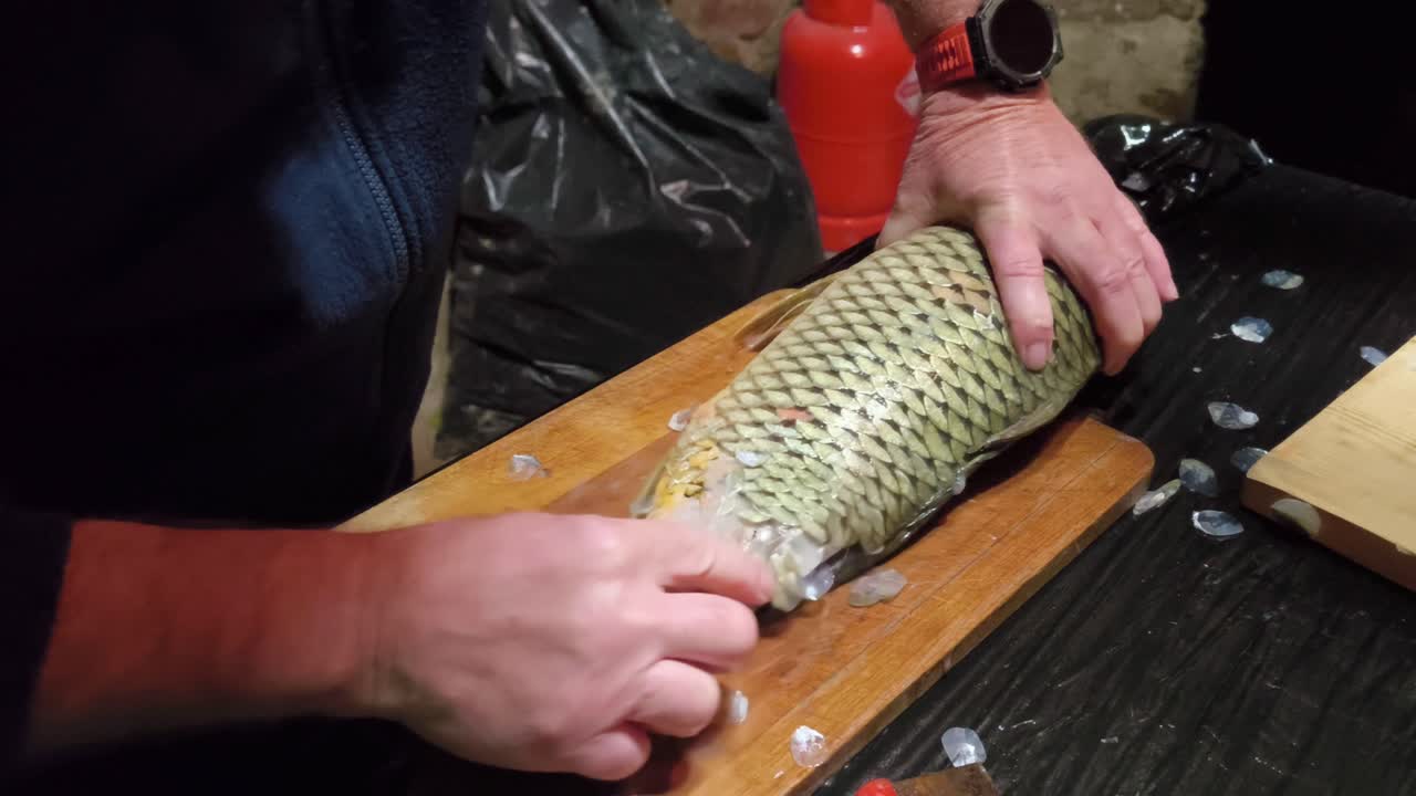 Hands scrape scales from a large fish on a wooden cutting board, removing them with firm strokes in a clear and realistic food preparation moment