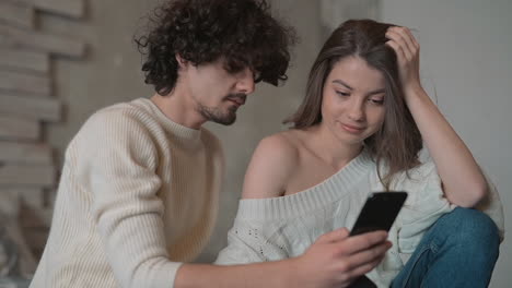 Young-Cute-Playful-Couple-Sitting-In-Bed-Looking-Something-Funny-On-The-Phone-And-Wearing-Winter-Clothes