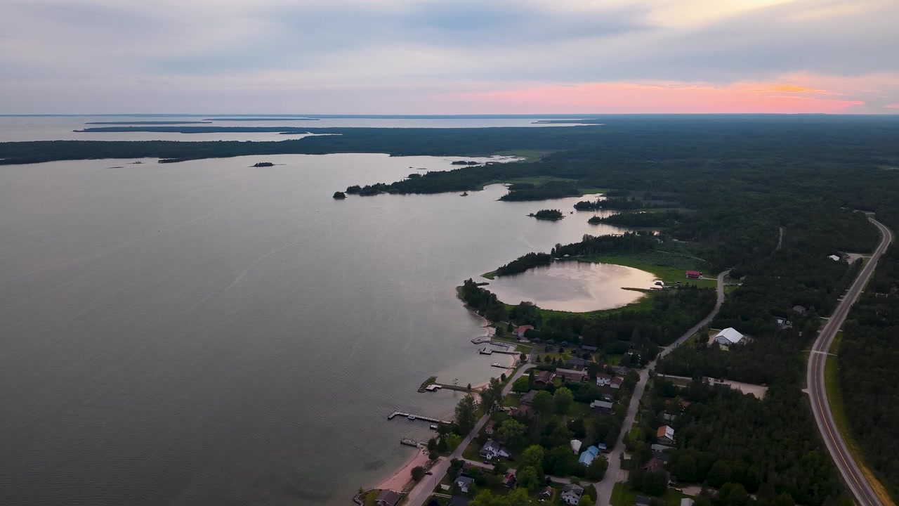 Panorama aereo dall'alto si affaccia su una piccola comunità sul lungolago del Lago Huron, con strada, foresta e isole, sotto un cielo al tramonto colorato