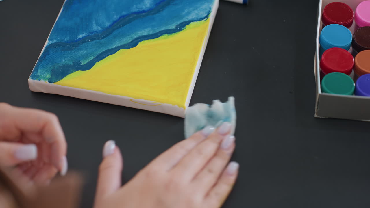 페인트 세트 근처의 캔버스 모서리를 닦는 클로즈업된 손, 보이는 푸른 물결과 노란 하늘 구도, 깔끔한 검은색 테이블 상판, 조심스러운 준비 동작, 정돈된 스튜디오 용품, 차분한 창작 루틴