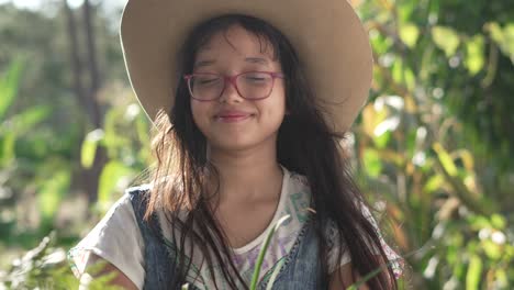 smiling girl wearing a hat and eyeglasses, standing in the middle of a botanical garden