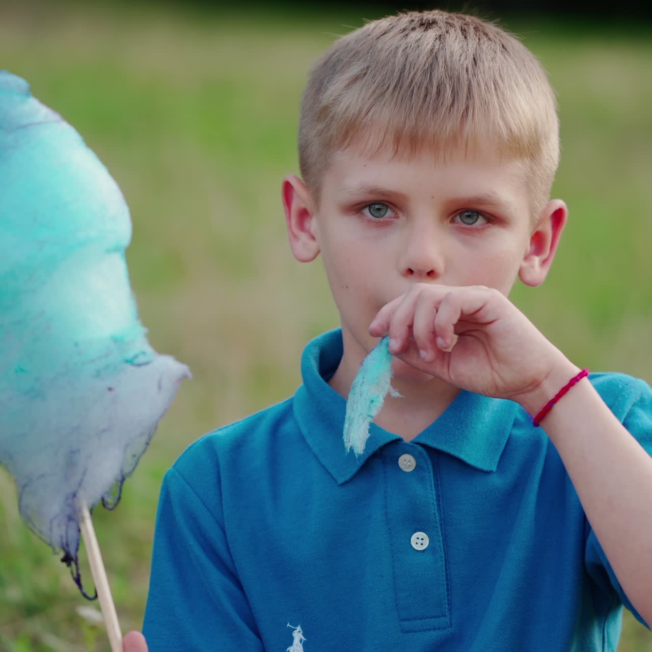 Portrait of a boy eating cotton candy. Cute boy in blue t-shirt sitting on the green grass and eats sweet candy-floss outdoors.