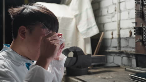 chinese student in white lab coat prepares for work by picking up protective glasses and a tablet in an automotive repair workshop, emphasizing safety and modern technology in engineering settings