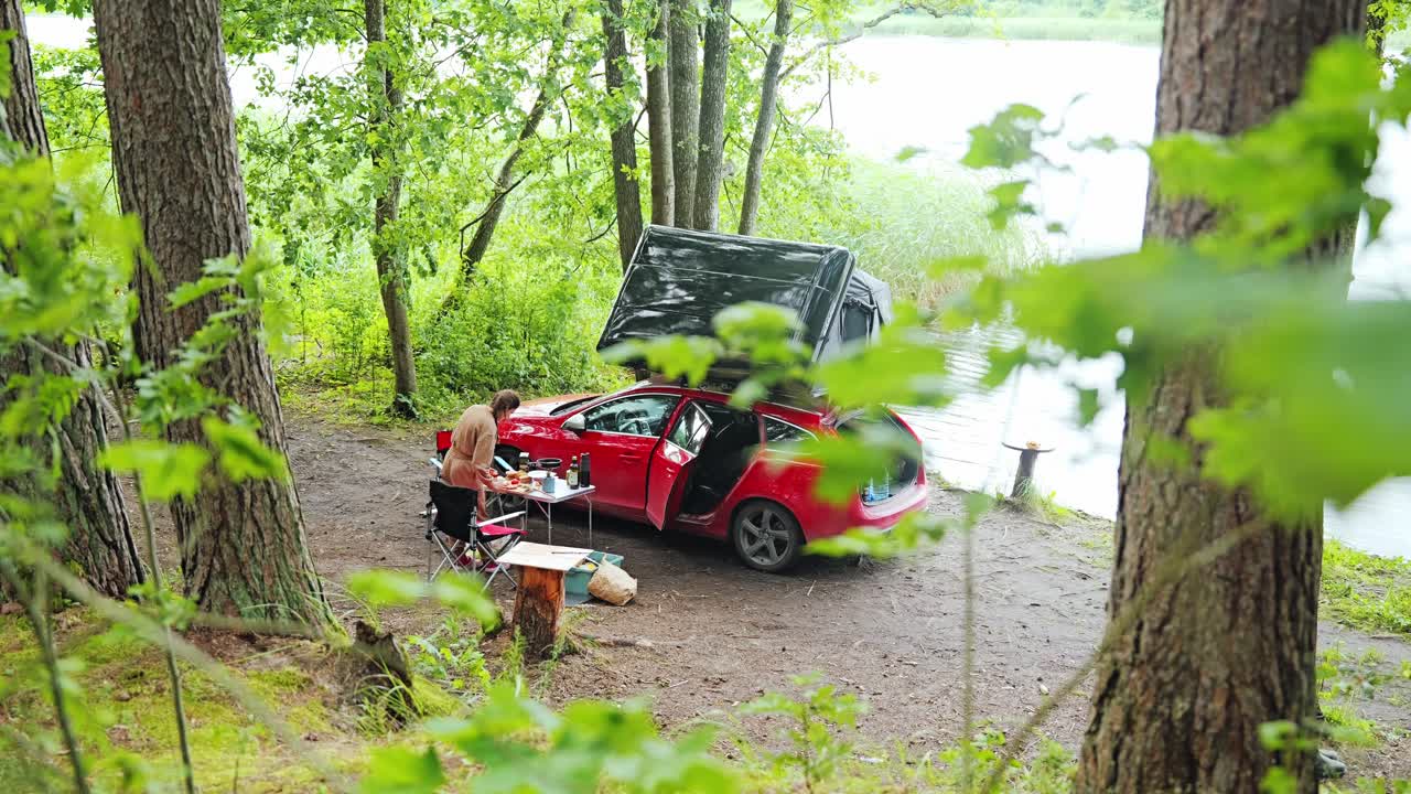 Peaceful solo camping by forest lake as woman cooks beside red rooftop car