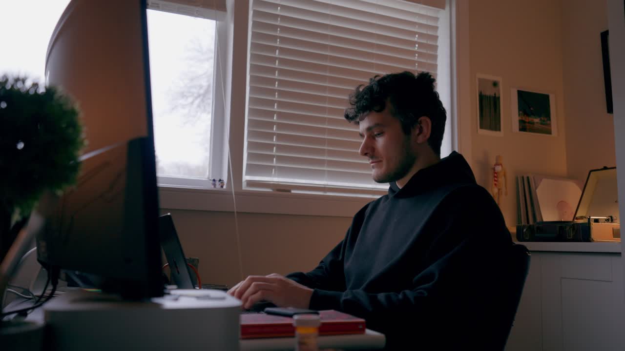 Vista laterale statica di un uomo che digita su una tastiera mentre lavora al computer in un ufficio domestico con una luce soffusa proveniente dalla finestra e un'illuminazione interna calda