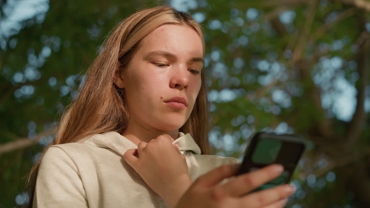 mujer joven con ropa casual opera atentamente el teléfono mientras sostiene su collar de capucha, la cara iluminada por la luz del sol filtrando a través de las hojas, el fondo borroso con vegetación