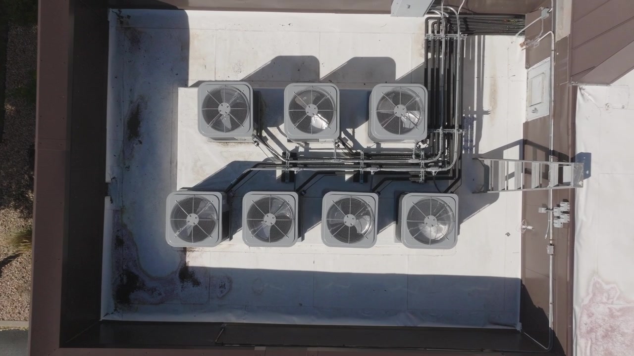 Riprese drone che mostrano unità di condizionamento sul tetto di un edificio commerciale, evidenziando sistemi HVAC industriali e architettura urbana sotto cieli sereni.