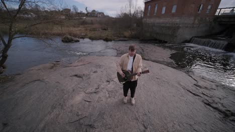 young man playing guitar sitting on the bank of a mountain river on a background of rocks. concept of freedom relaxation. place