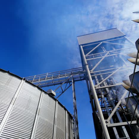 Huge hangar premises. Grain elevators outdoors. White vapor released from metal granary for drying grains. Close-up. View from below.