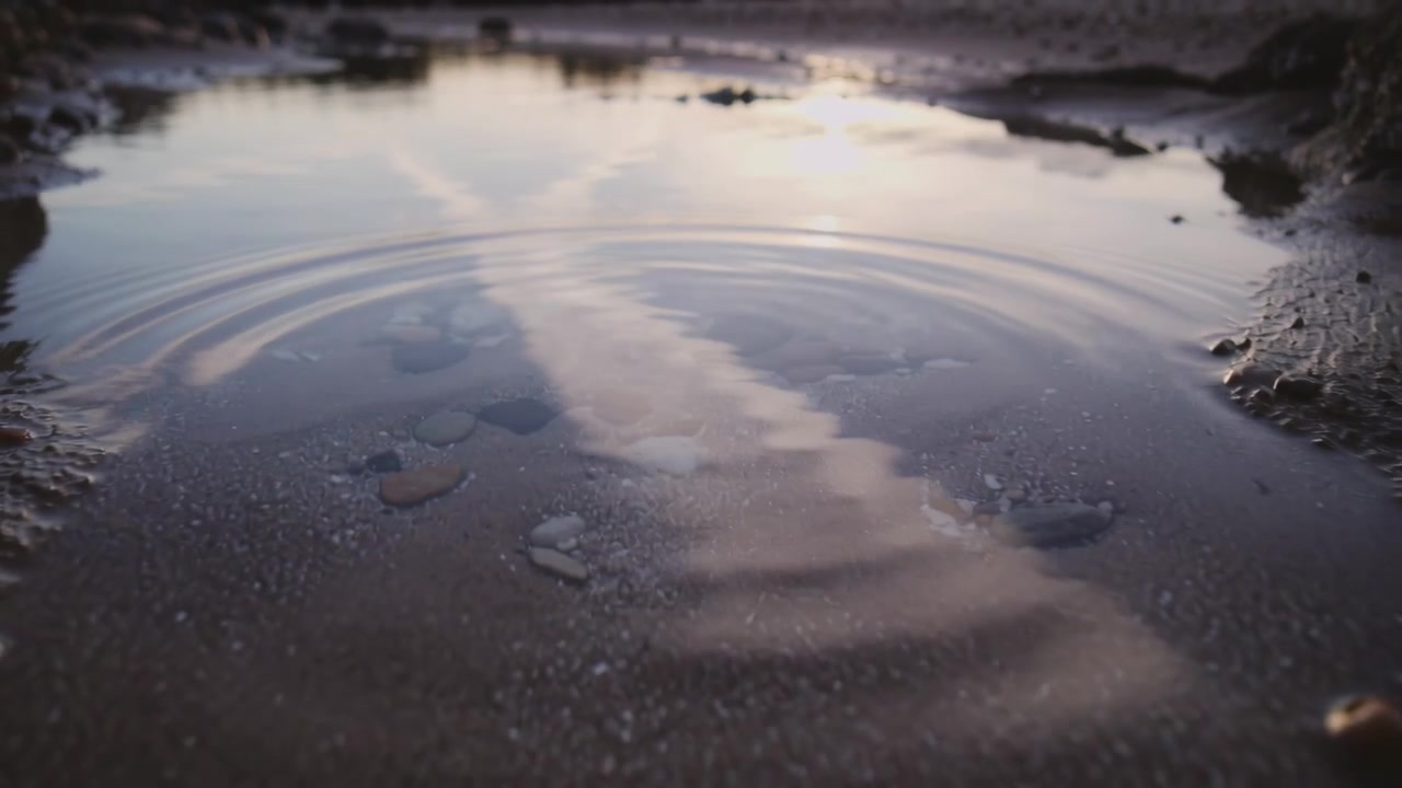 Rippende tidevannskulp som skaper runde ringer ved sandstranden etter at en stein traff vannet, reflekterer kondensstripe