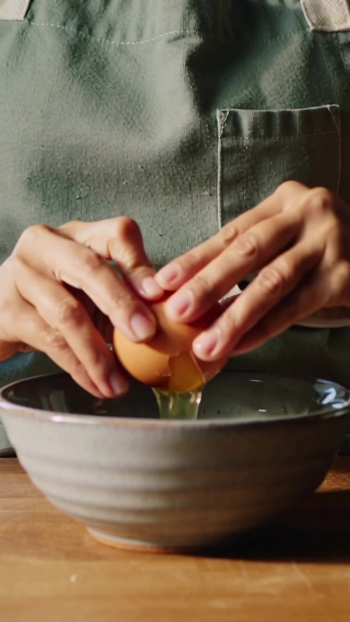 Vertical video: Cracking cook in sage apron looking, opening brown egg into bowl in kitchen mixing
