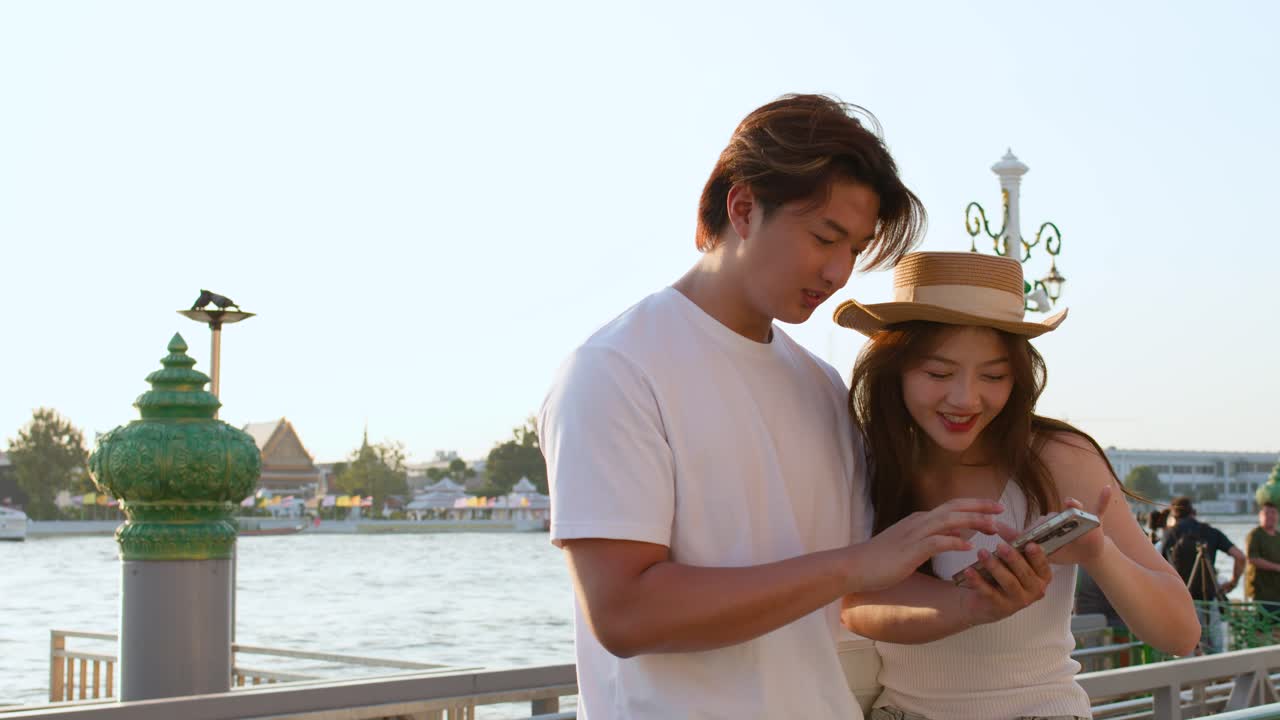 Smiling couple interacts with smartphone outdoors, warm sunset lighting, riverside temple background, handheld camera