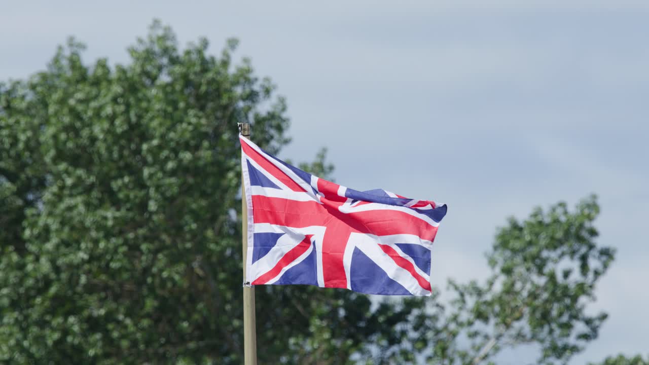 Union flag flutters vigorously on flagpole outdoors, bright daylight, steady camera, leafy background, Cromarty