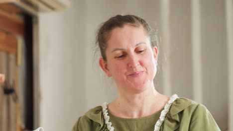 portrait view of cheerful chef in green dress smiling joyfully while holding electric mixer coated with thick whipped dough and lifting empty bowl upside down to show stickiness