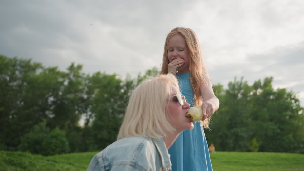 young girl giving mom pear while eating snack with other hand during picnic in park, denim jacket mother, warm light, playful sharing between parent and child on green grass