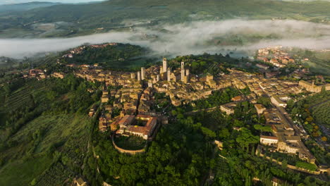 fly over the middle-aged town of san gimignano surrounded by green hills on a foggy sunrise in tuscany, italy