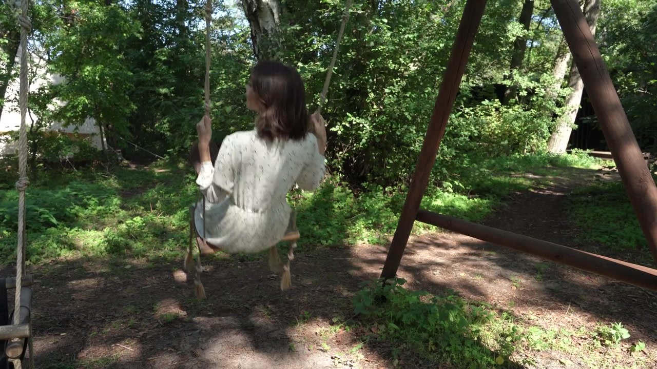 Ragazza con capelli scuri in un vestito campestre bianco si dondola su un'altalena di legno nel bosco.
