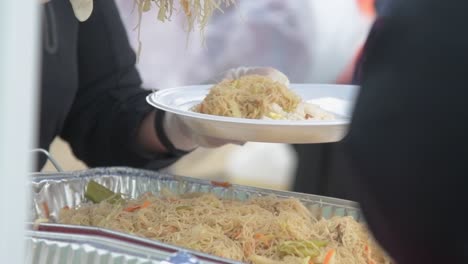 person serving fresh hot noodles on white plastic plate with tongs, outdoor buffet