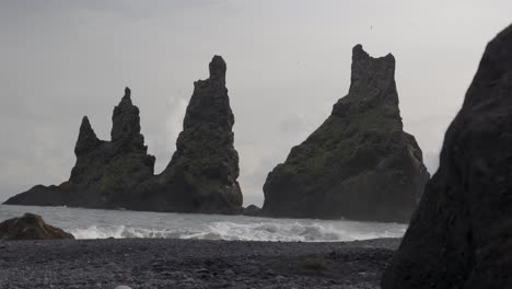 Hermosas-Formaciones-Rocosas-De-Lava-En-Un-Paisaje-Cambiante-Y-Brumoso,-Olas-Chapoteando-Contra-La-Playa-De-Arena-Negra